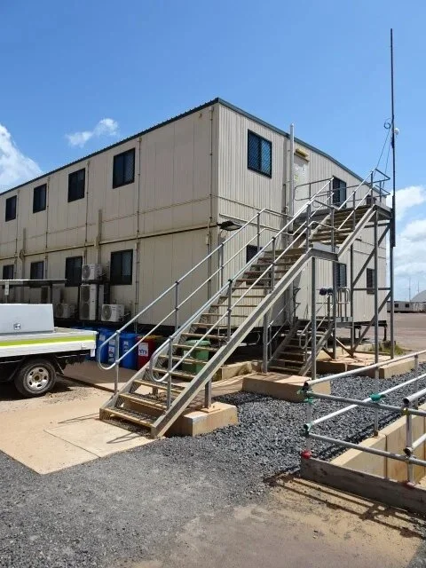 A two-story portable building with metal stairs and handrails leading to the upper level, surrounded by air conditioning units, vehicles, and drums, under a clear, sunny sky.