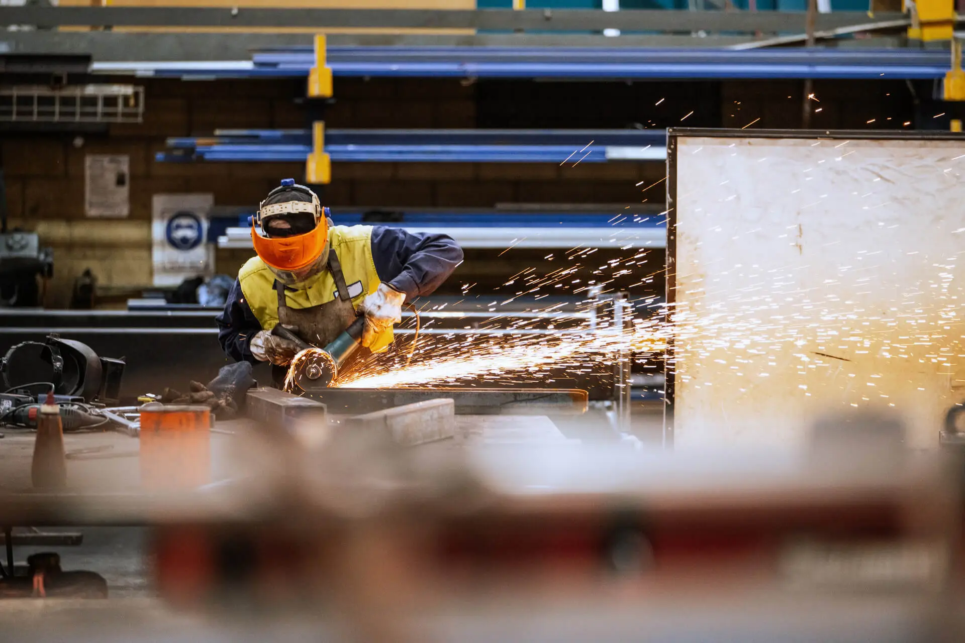 A worker in safety gear uses an angle grinder on metal, creating bright sparks in an industrial workshop filled with shelves, equipment, and metal materials.