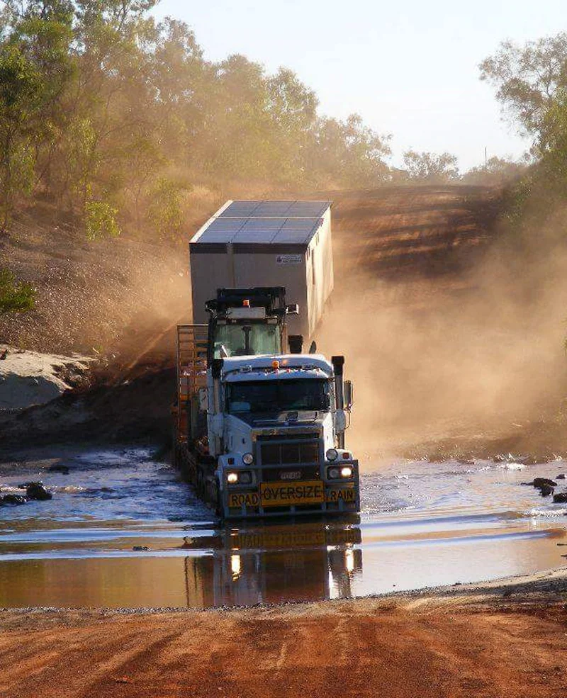 A large road train truck crosses a shallow, muddy waterhole on a dirt road surrounded by trees, kicking up dust as it drives through the rugged, remote landscape.