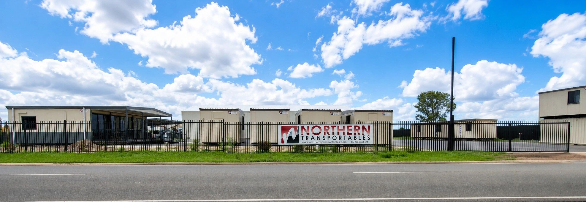 A fenced commercial property with modular buildings, a large sign reading "Northern Transportables" on the fence, and a partly cloudy sky above.