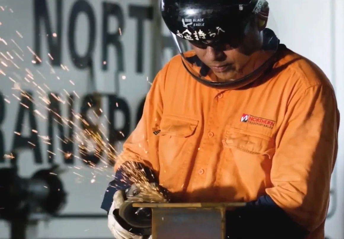 A worker in an orange safety shirt and helmet uses a grinder, sending sparks flying as he works on a metal piece indoors. The background features large, partially visible, black text on a white wall.