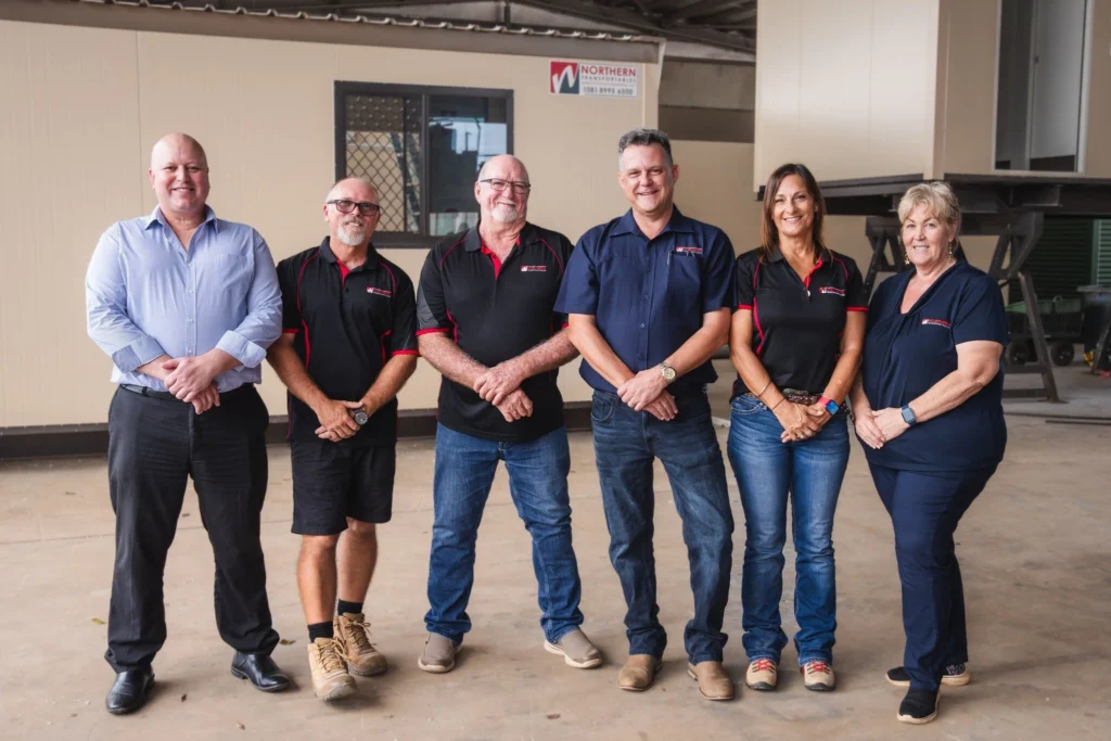 Six adults stand in a row, smiling, in front of a building with a "Northern" sign. Five wear branded work shirts and casual pants; one man is in business attire. The setting appears to be an industrial or workshop area.