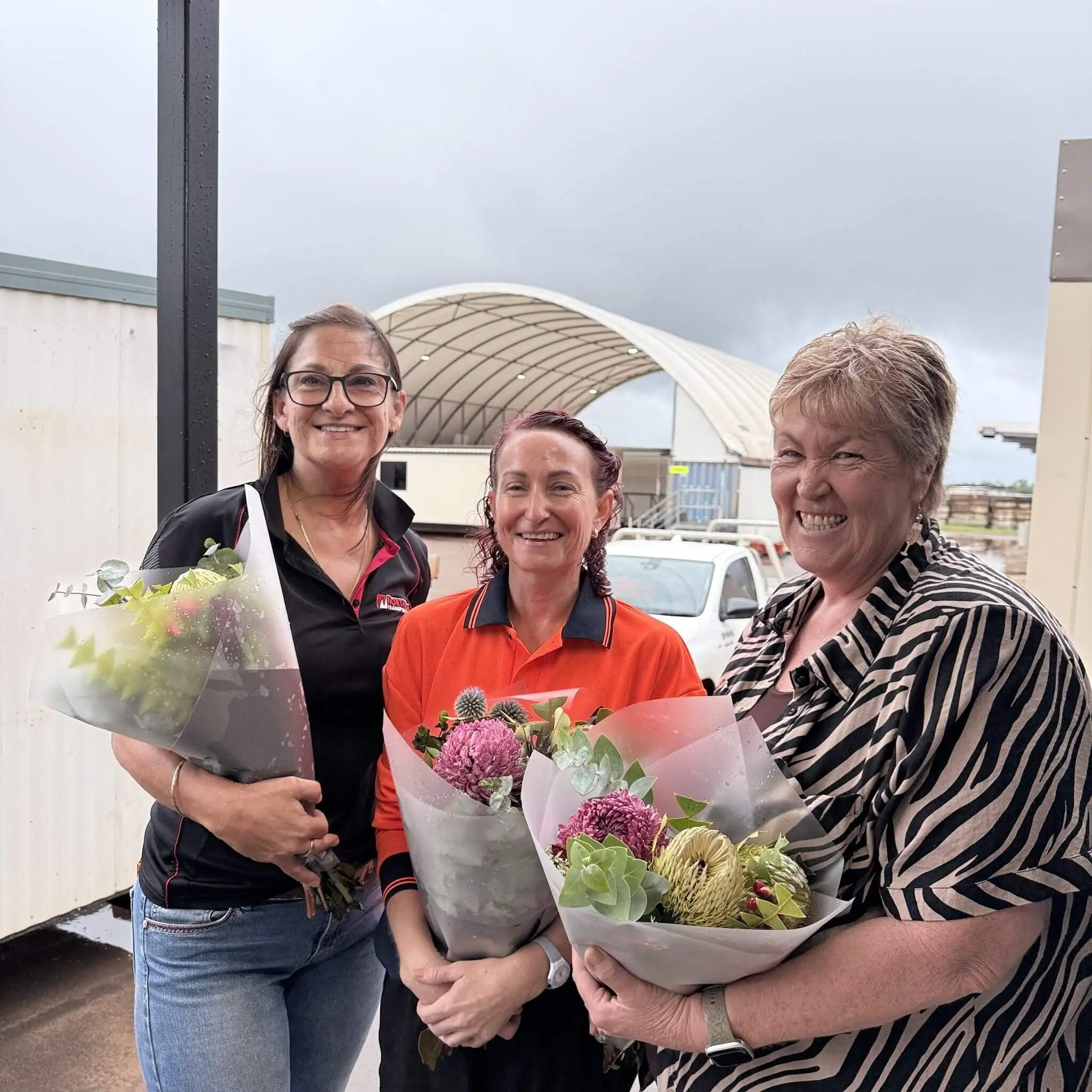 Three smiling women stand outside holding bouquets of flowers. Two wear casual shirts, one in an orange work shirt. A white vehicle and a curved metal shelter are visible in the background under a cloudy sky.