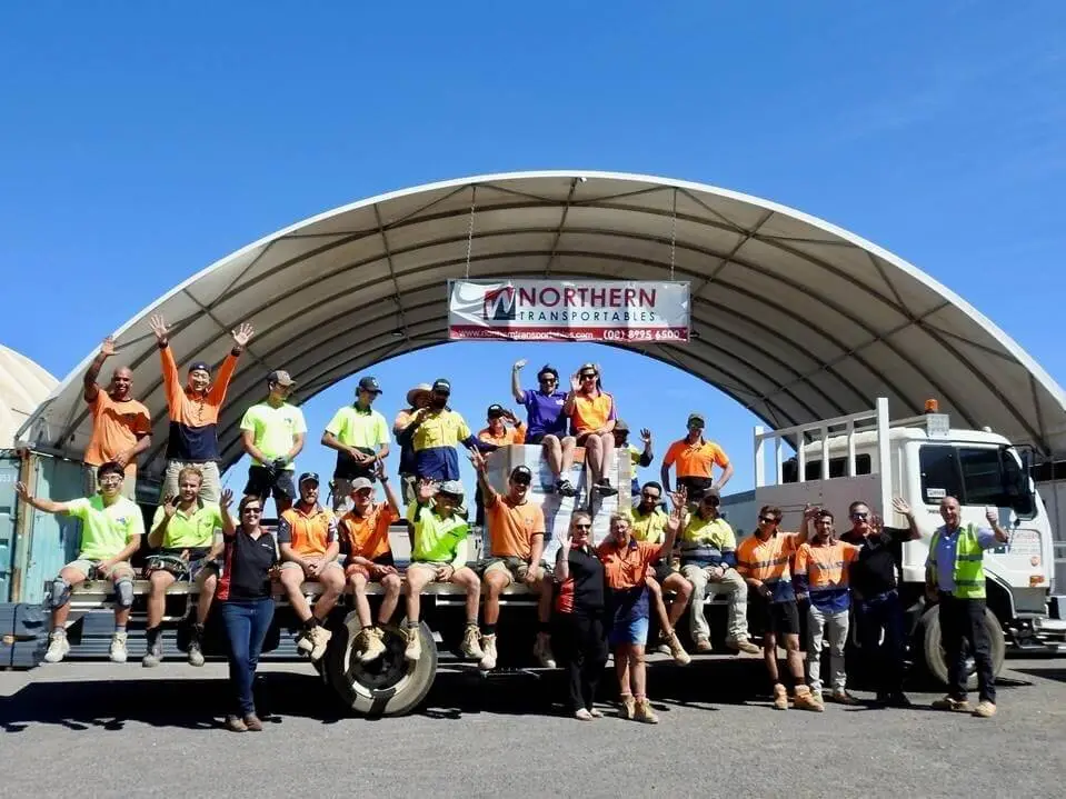 A large group of construction workers in high-visibility clothing pose, smiling, on and in front of a flatbed truck under a large arched shelter with a "Northern Transportables" banner.