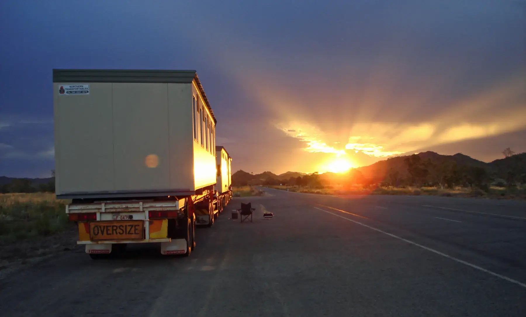 A large truck with an "OVERSIZE" load sign is parked on the roadside at sunset. The sun is setting behind distant hills, casting golden rays and illuminating the sky with a warm glow.