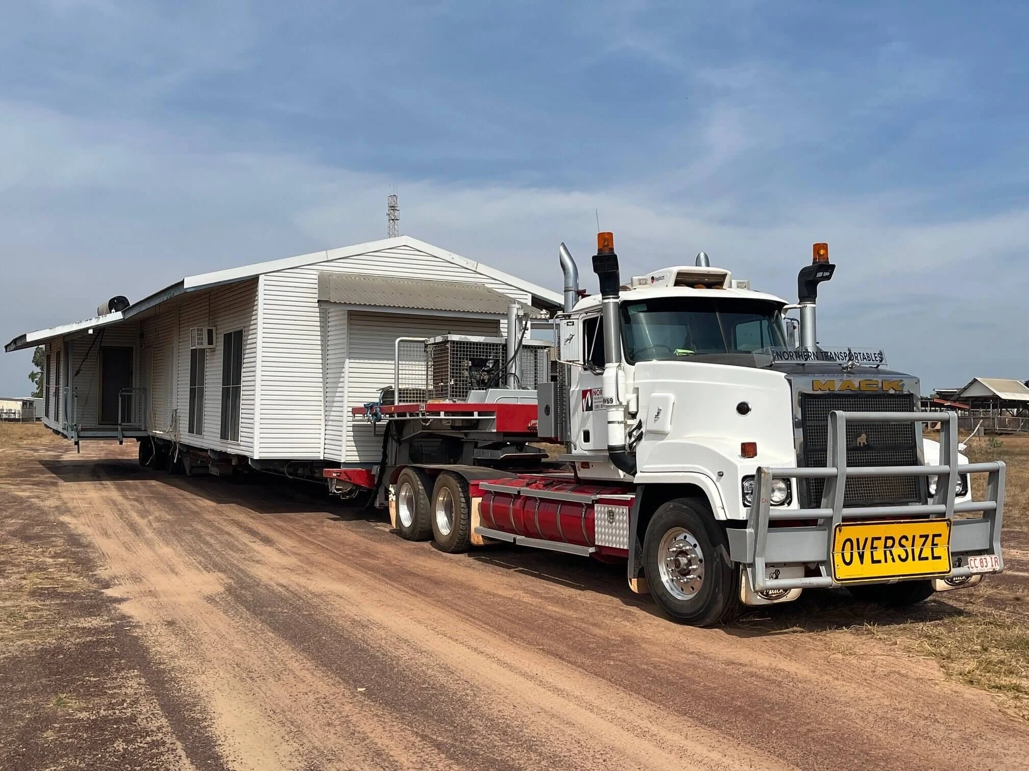 A large white truck labeled "OVERSIZE" transports a single-story white house along a dirt road under a blue sky. The house is securely loaded on the trailer behind the truck.