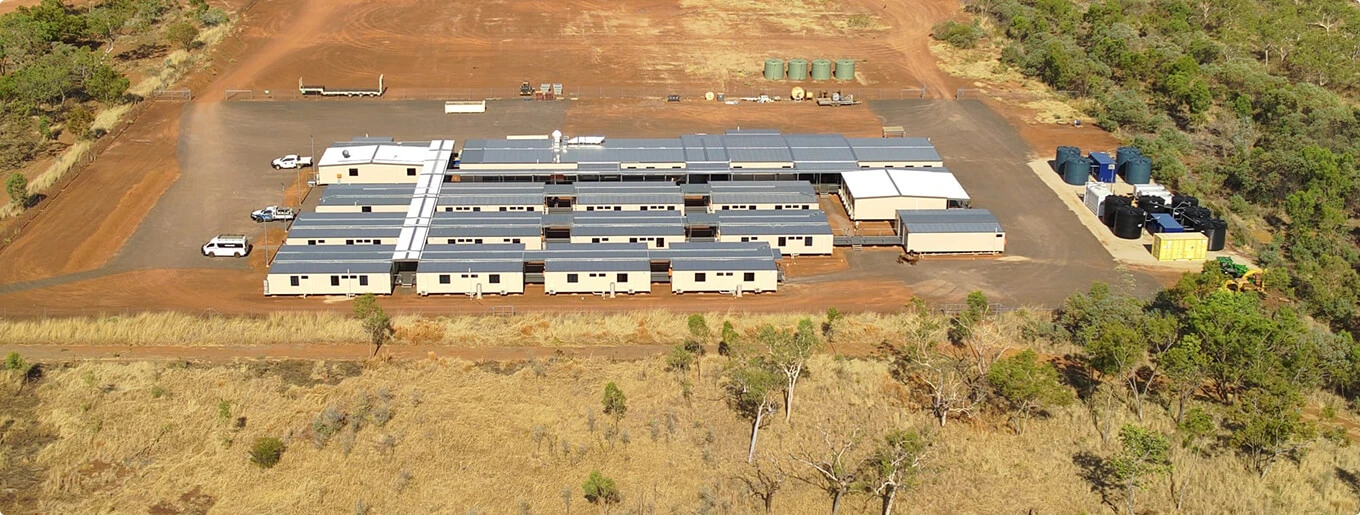 Aerial view of a remote, modular building complex with several white structures, parked vehicles, water tanks, and surrounding red earth and sparse vegetation.