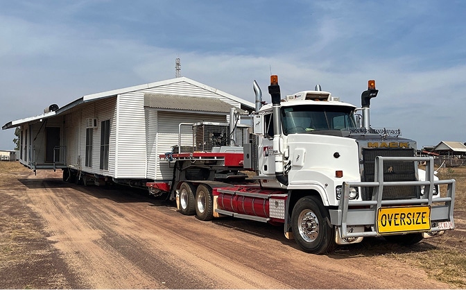A large white truck with an "OVERSIZE" sign transports a house on a trailer along a dirt road under a clear sky.