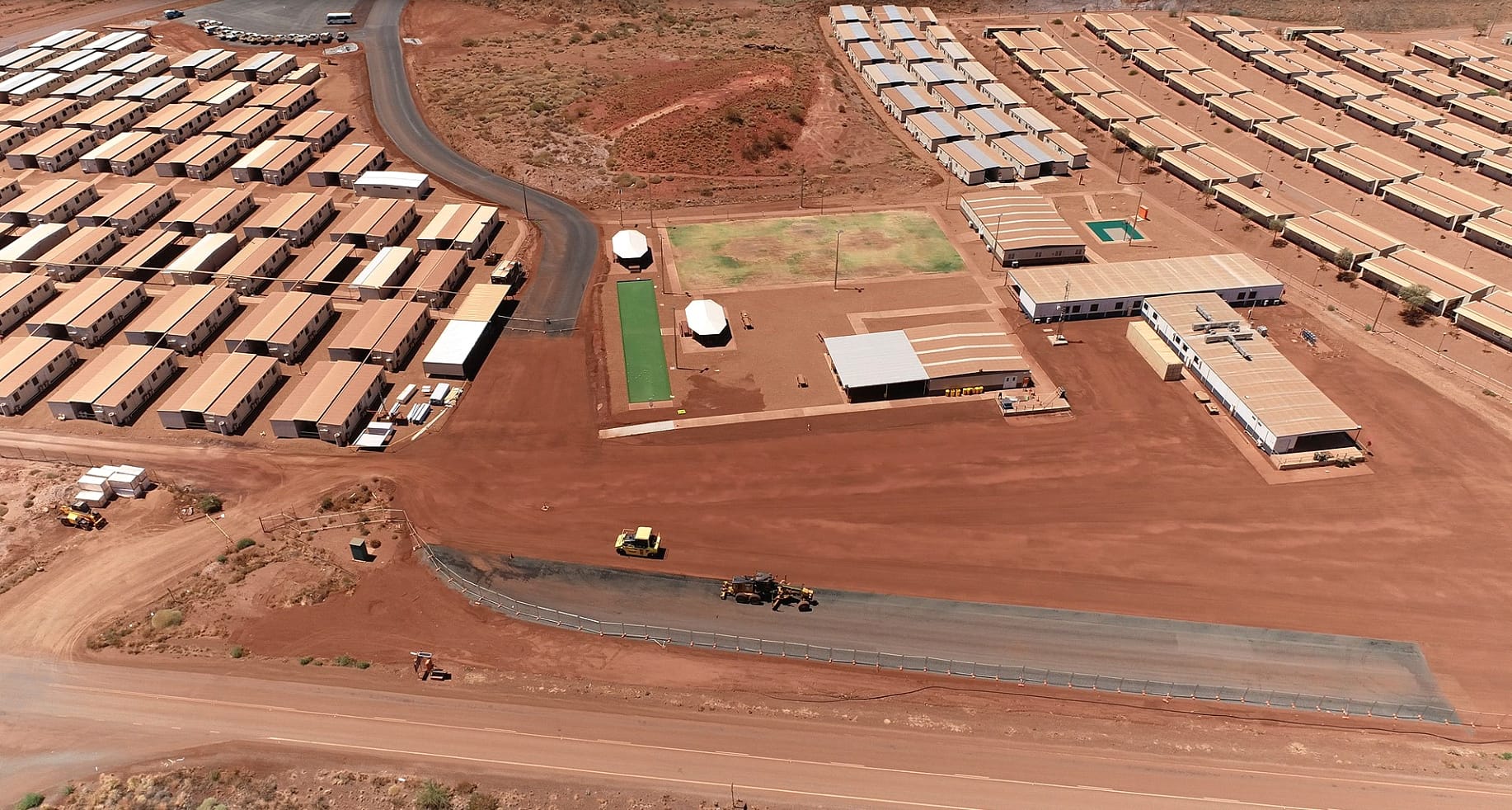 Aerial view of a remote desert facility with rows of portable buildings on reddish soil, a few vehicles, grassy areas, and paved roads, surrounded by barren, rocky landscape.