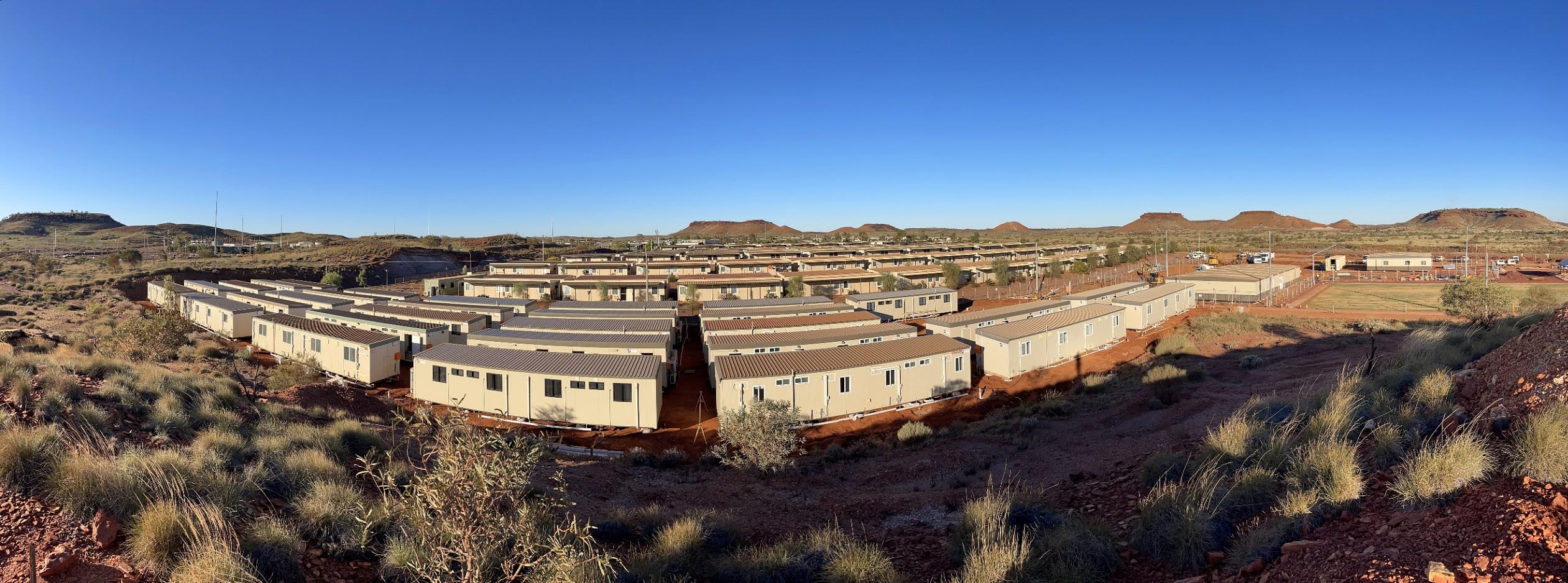 Rows of beige modular buildings are arranged in a remote, arid landscape with red soil, sparse vegetation, and distant low hills under a clear blue sky.