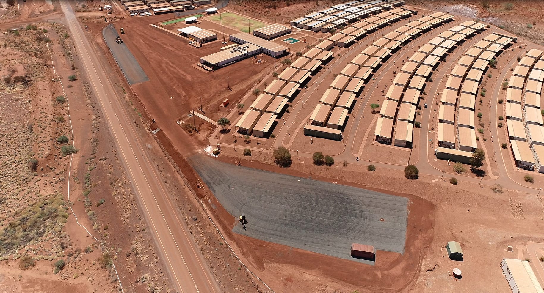 Aerial view of a remote, desert-like area with rows of long, rectangular buildings, possibly housing or dormitories, next to a paved road and sports facilities, surrounded by reddish-brown terrain.