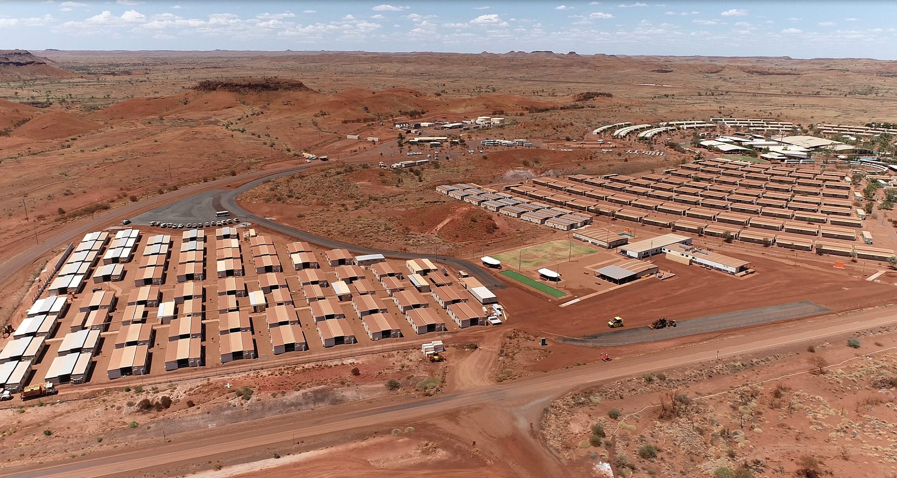 Aerial view of a large mining camp in a red, arid landscape, featuring rows of modular buildings, access roads, and sparse vegetation, with distant hills under a partly cloudy sky.