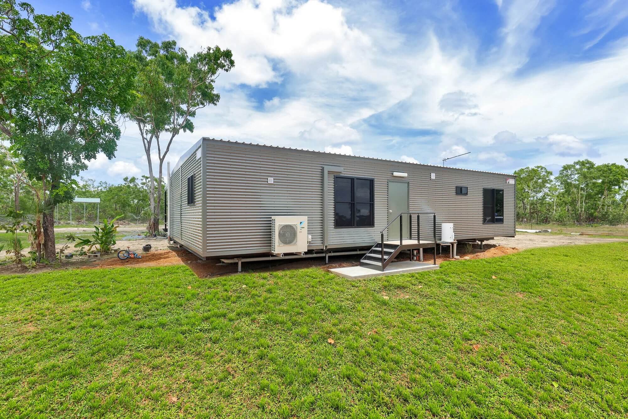 A modern, single-story, prefabricated home with gray metal siding and several windows sits on a grassy lot with trees and a blue sky in the background. Steps lead to the entrance, and an air conditioning unit is installed outside.