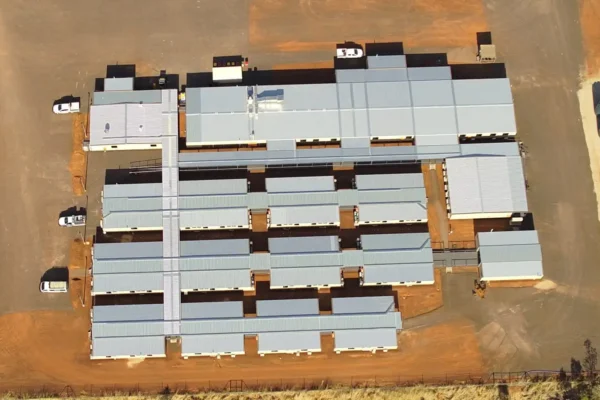 Aerial view of a large, rectangular complex with multiple connected gray-roofed buildings, parked vehicles, and surrounding reddish-brown dirt. Grass or vegetation borders the lower edge of the facility.