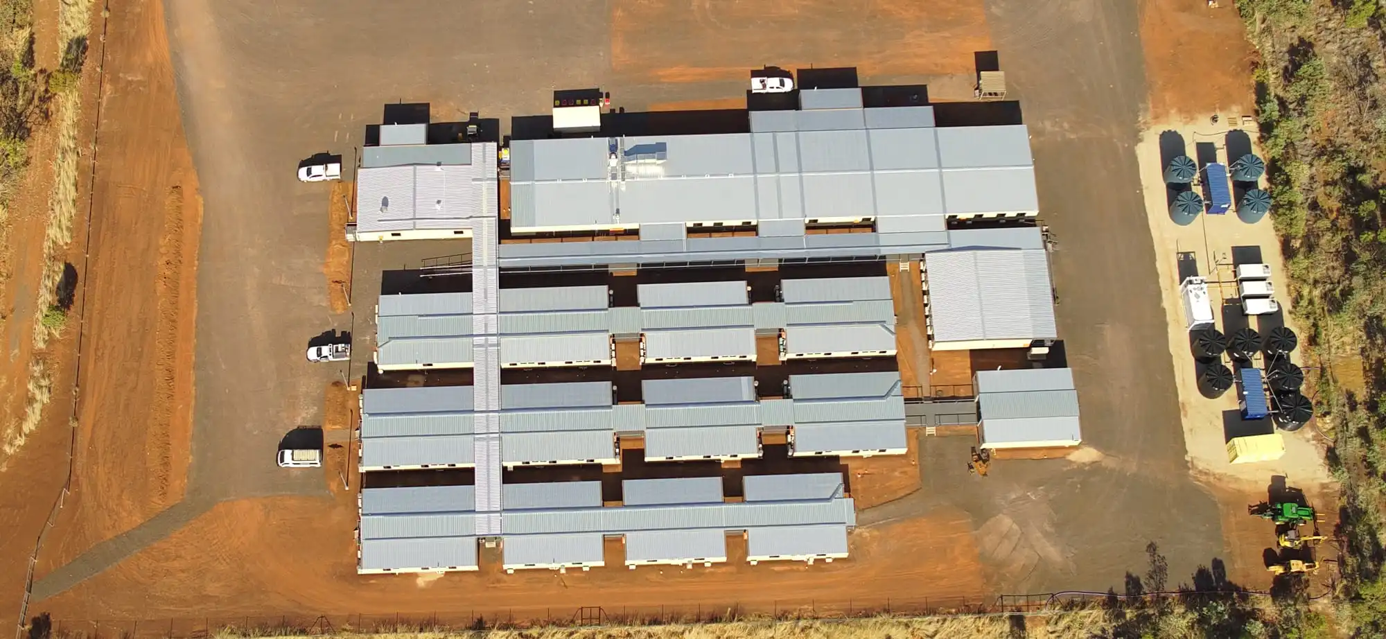 Aerial view of a large, rectangular complex with multiple connected gray-roofed buildings, parked vehicles, and surrounding reddish-brown dirt. Grass or vegetation borders the lower edge of the facility.