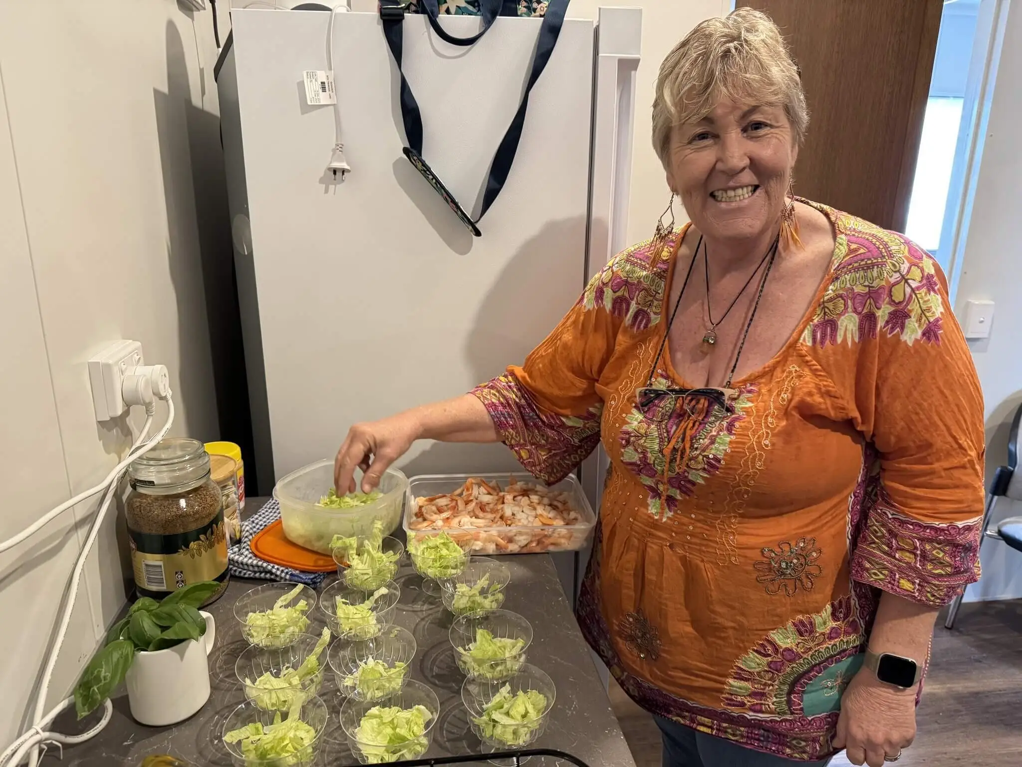 A smiling woman in an orange patterned top prepares food in a kitchen, placing lettuce into glass bowls. A tray of shrimp, glass jars, and other ingredients are on the counter beside her.