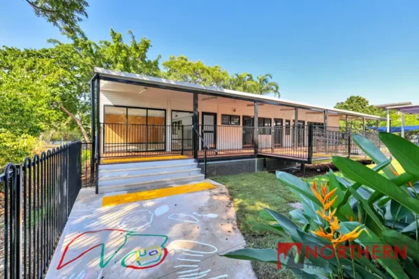 A modern, single-story building with a ramp and stairs leading to a covered veranda, surrounded by trees. Colorful playground markings are on the concrete, and a bright bird of paradise plant is in the foreground.