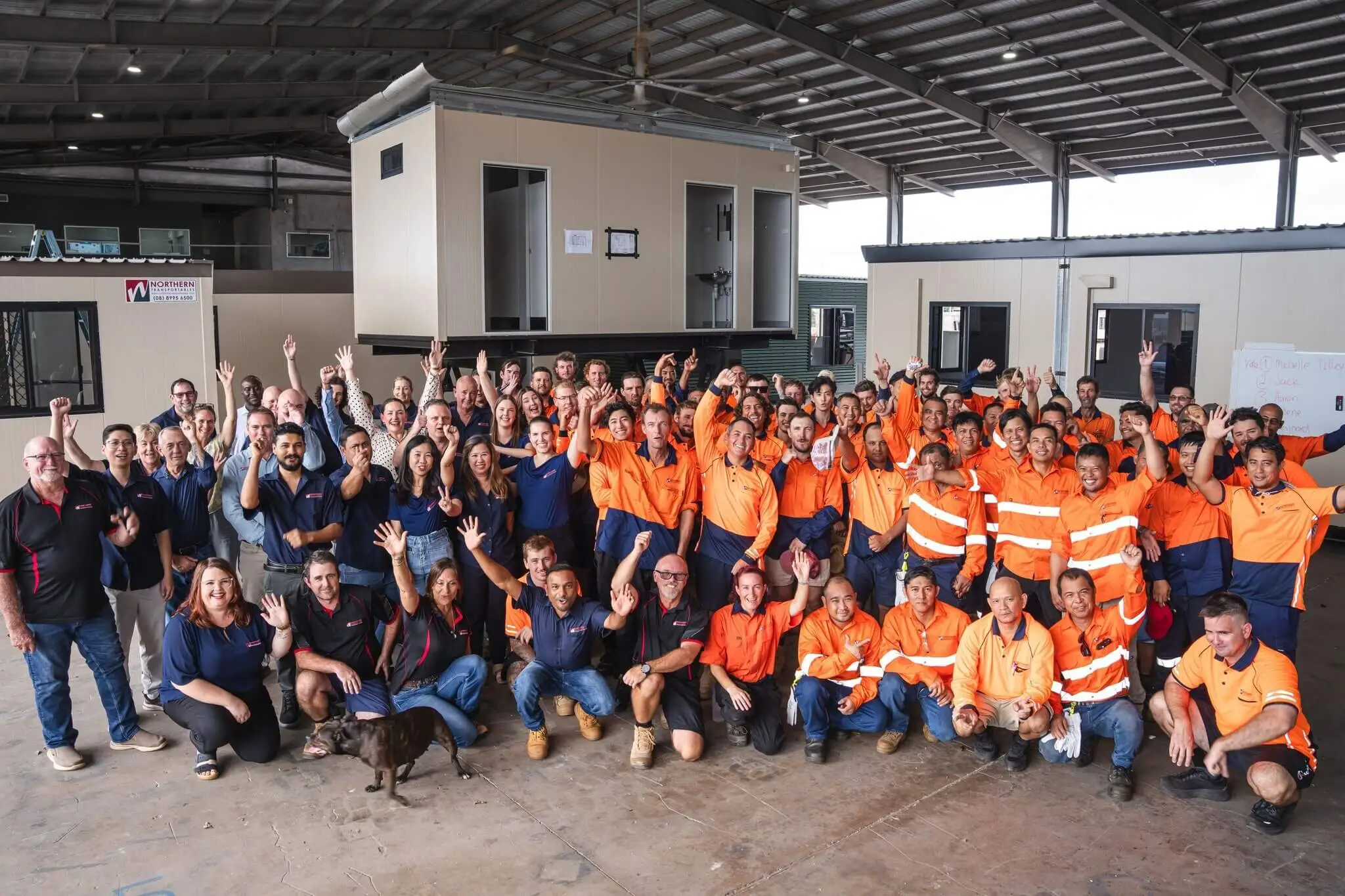 A large group of workers, many wearing orange high-visibility clothing, pose together and smile inside a warehouse. Some people are raising their arms in celebration. Modular buildings are visible in the background.
