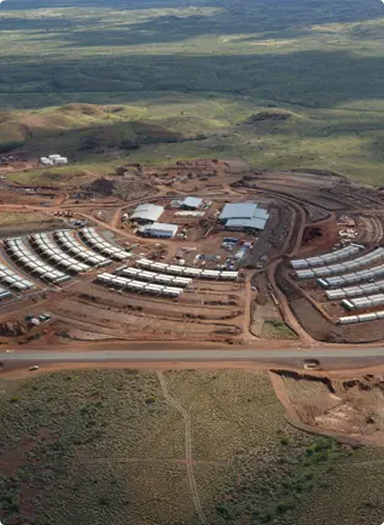 Aerial view of a construction site with curved rows of temporary white structures, buildings, roads, and vehicles, surrounded by green and brown landscape.